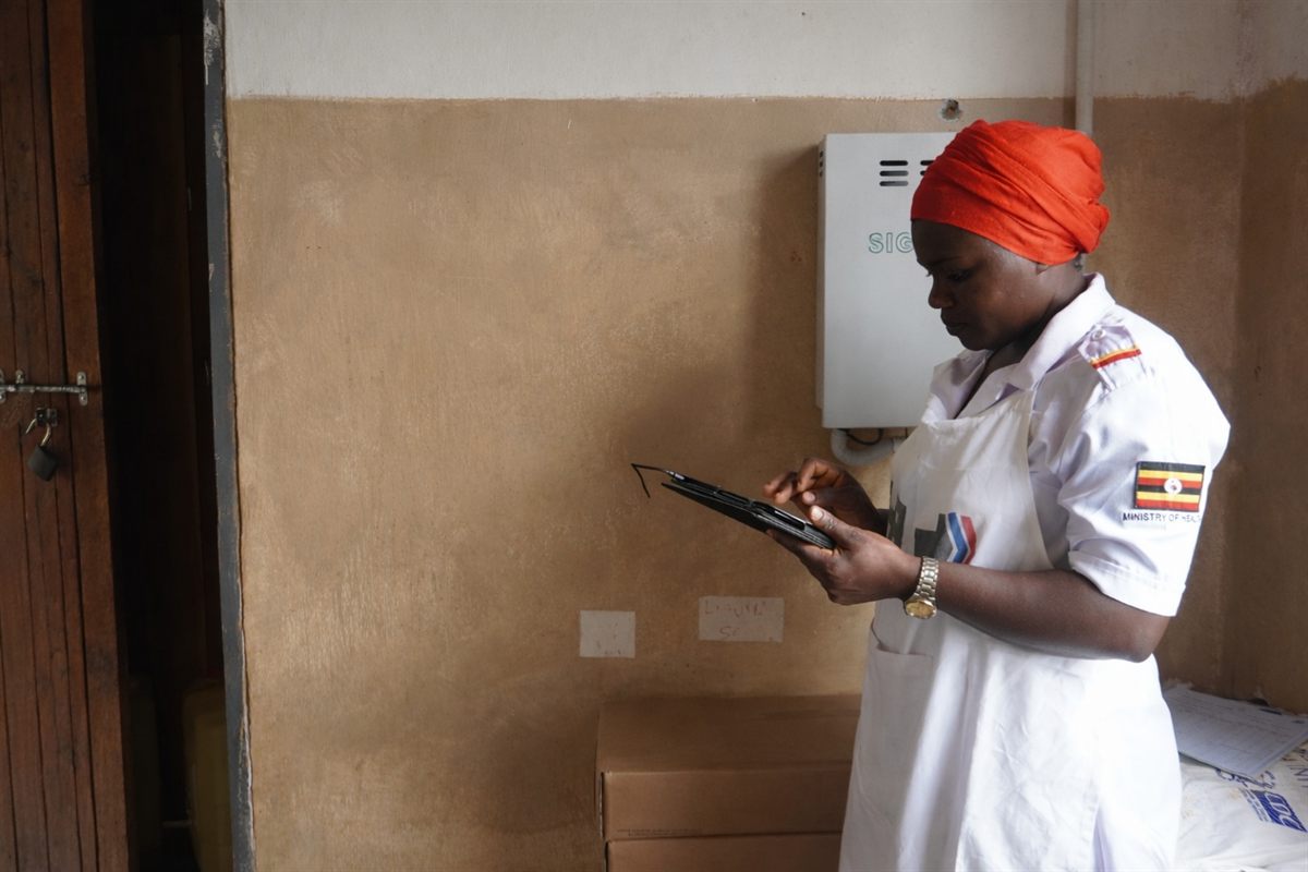 A health worker in Ministry of Health uniform entering records on a tablet, beside a wall-mounted S+ Platform enclosure.