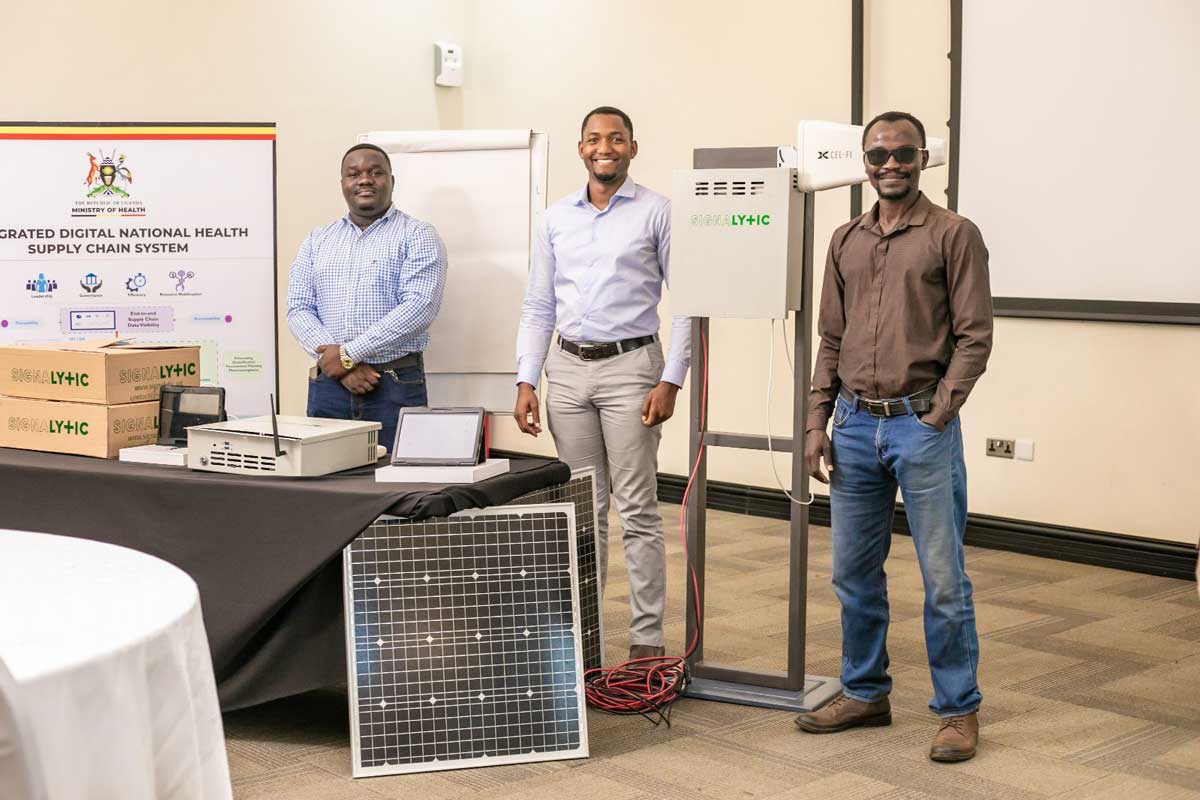 Three men with Signalytic equipment — a solar panel, an S+ Platform enclosure and a Cel-Fi antenna — beside a Uganda Ministry of Health supply chain banner.